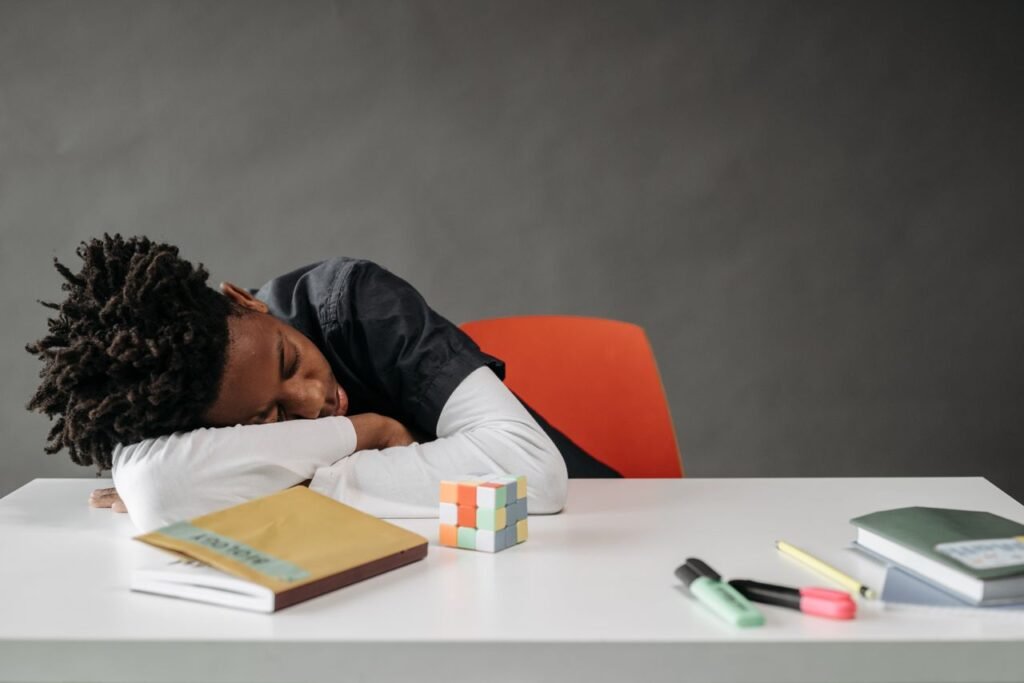 A young boy sleeping at a desk with books and a Rubik's cube, looking weary.